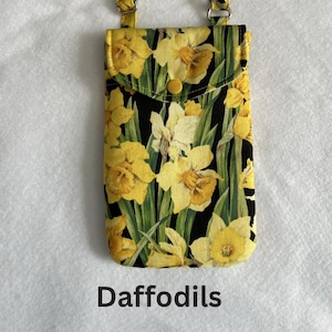 May include: A yellow floral print phone case with a flap closure and a button. The case features a daffodil pattern on a black background, with green leaves. The word "Daffodils" is printed at the bottom.