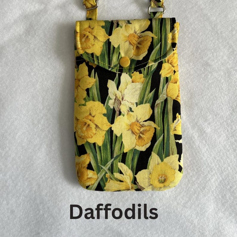 May include: A yellow floral print phone case with a flap closure and a button. The case features a daffodil pattern on a black background, with green leaves. The word "Daffodils" is printed at the bottom.