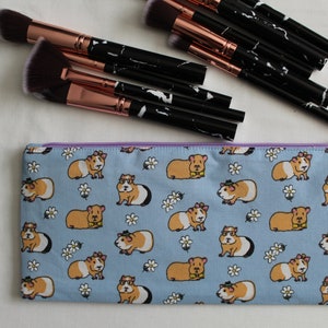 Puede incluir: Una bolsa de maquillaje con cremallera de color azul claro con un patr&oacute;n de conejillos de indias con gafas y pajaritas sobre un fondo blanco con peque&ntilde;as flores blancas.