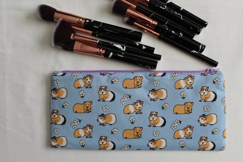 Puede incluir: Una bolsa de maquillaje con cremallera de color azul claro con un patr&oacute;n de conejillos de indias con gafas y pajaritas sobre un fondo blanco con peque&ntilde;as flores blancas.