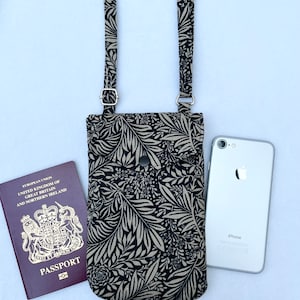 May include: A black and white floral patterned crossbody phone bag with a silver snap closure. The bag is shown with a passport and a white iPhone.