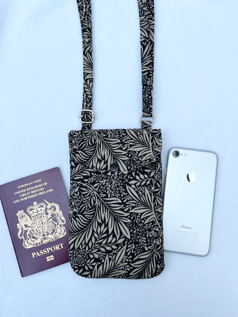 May include: A black and white floral patterned crossbody phone bag with a silver snap closure. The bag is shown with a passport and a white iPhone.
