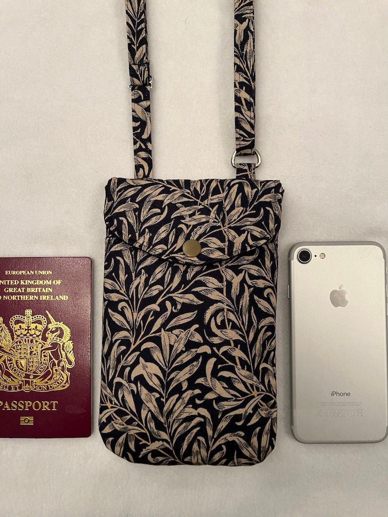 May include: A black and cream floral patterned phone pouch with a small pocket on the front. The pouch has a long adjustable strap and is shown next to a passport and an iPhone.