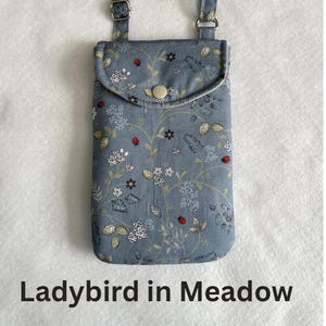 May include: A blue floral print phone bag with a flap closure and adjustable strap. The bag features a pattern of white flowers, green leaves, and red ladybugs. The text "Ladybird in Meadow" is printed at the bottom.