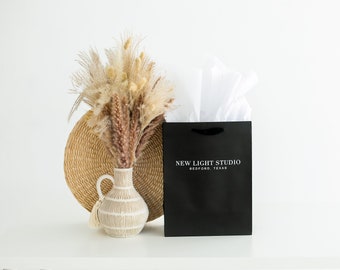 May include: A black gift bag with the text "NEW LIGHT STUDIO BEDFORD, TEXAS" is filled with white tissue paper. A vase with dried flowers and a woven placemat are to the left of the bag, all on a white table.