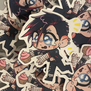 May include: A selection of stickers depicting a cartoon character with black hair, red highlights, and bandages. The character is holding a cupcake with pink icing. The stickers have a white border and are spread across a surface.