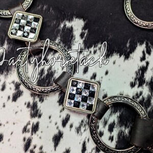 May include: Close-up of horse tack featuring silver-toned rings and square accents with a black and white checkered pattern. The leather straps are dark brown. The text "Partyhorsetack" is written in a cursive font.