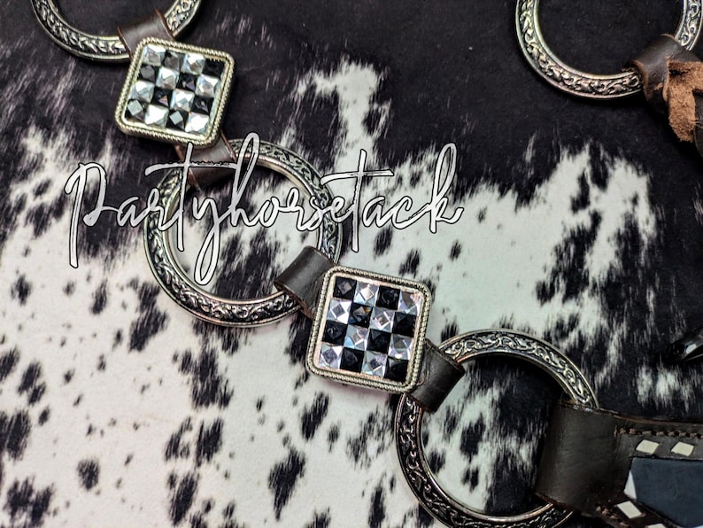 May include: Close-up of horse tack featuring silver-toned rings and square accents with a black and white checkered pattern. The leather straps are dark brown. The text "Partyhorsetack" is written in a cursive font.