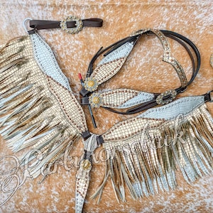 May include: A horse tack set featuring a gold and silver design with fringe. The set includes a bridle, breast collar, and a browband, all adorned with decorative buckles and studs. The items are arranged on a brown surface.