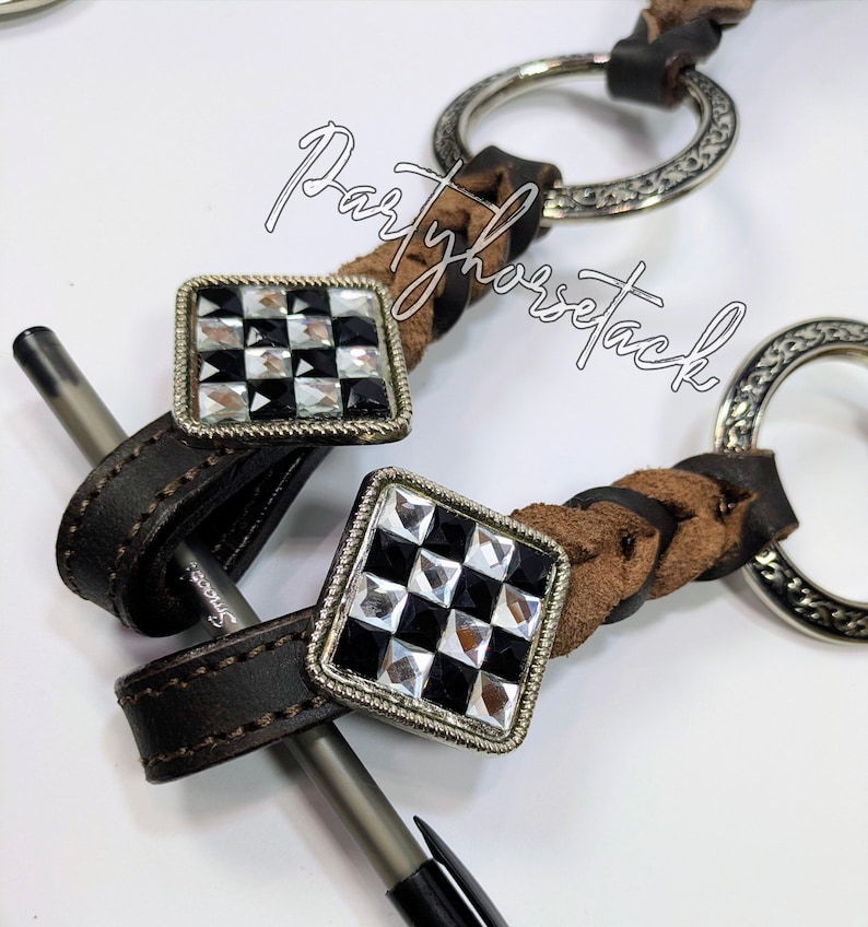 May include: Close-up of equestrian equipment featuring a dark brown leather strap with a silver metal ring and a decorative square with a black and white checkered pattern. The text "Partyhorsetack" is visible.