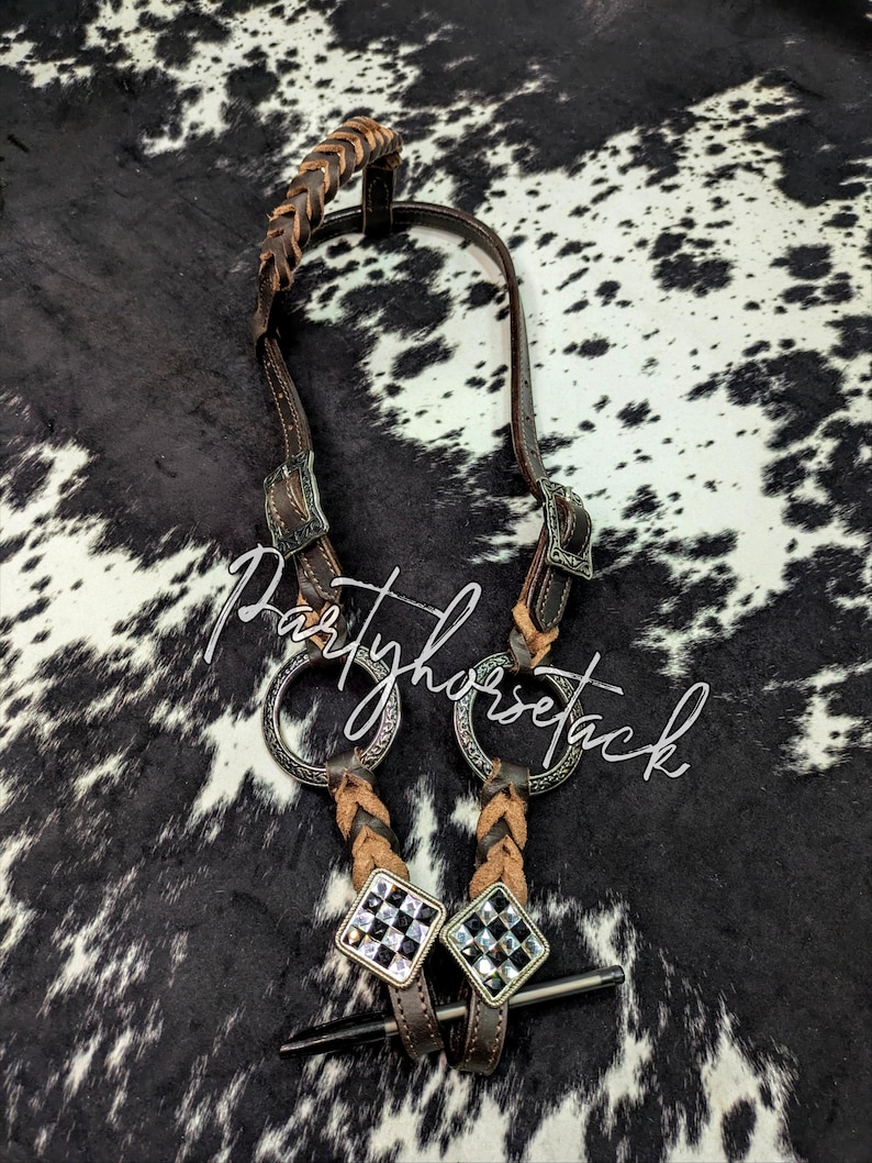 May include: A brown leather horse bridle with decorative silver-toned buckles and accents. The bridle features a braided leather headstall and cheek pieces, with two silver rings and a bit. The text "Partyhorsetack" is visible.