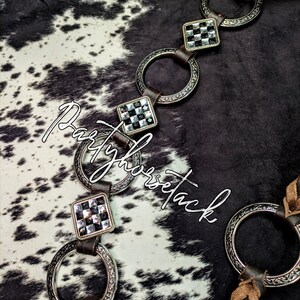 May include: Close-up of a horse bridle with silver-toned metal rings and square accents with black and clear stones. The bridle has brown leather straps and is set against a black and white cowhide pattern. The text "Partyhorsetack" is visible.
