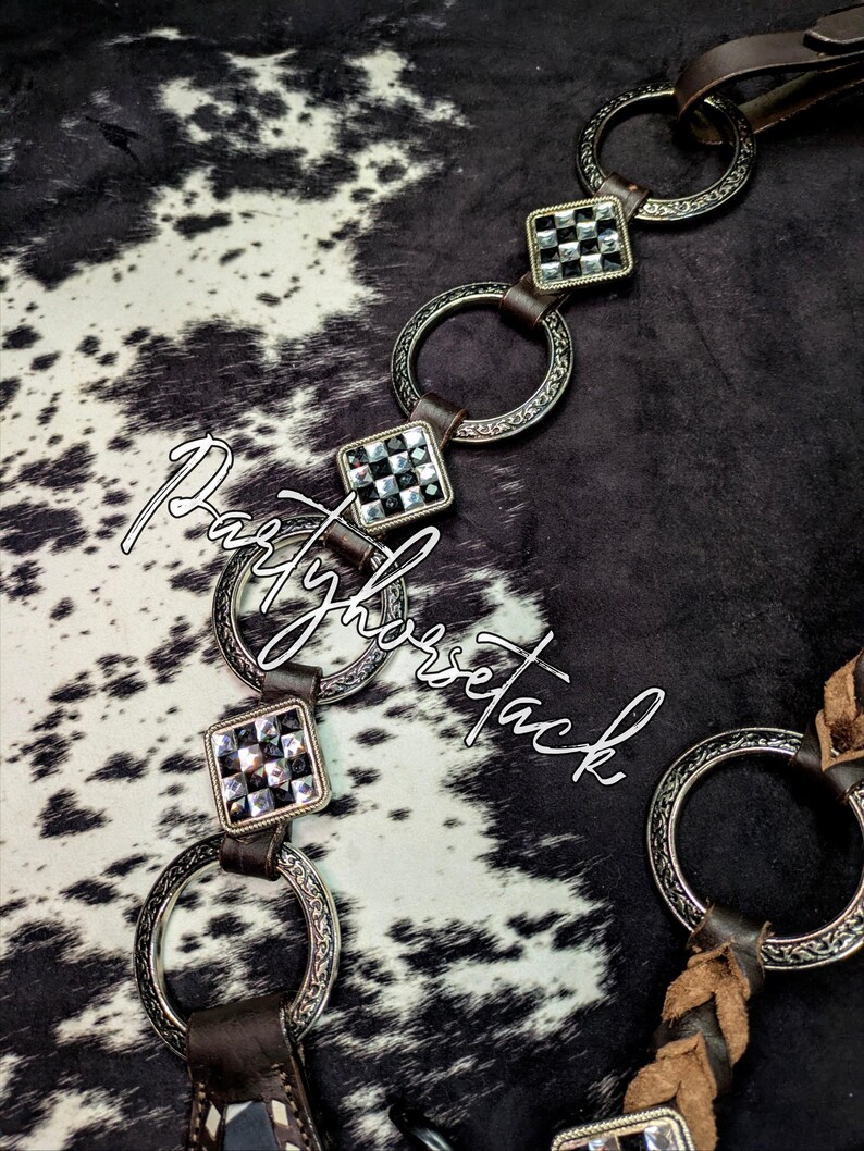May include: Close-up of a horse bridle with silver-toned metal rings and square accents with black and clear stones. The bridle has brown leather straps and is set against a black and white cowhide pattern. The text "Partyhorsetack" is visible.