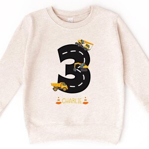 Peut inclure: Un sweat à capuche beige à manches longues avec le chiffre "3" noir sur le devant. Le chiffre est conçu pour ressembler à une route avec des véhicules de chantier jaunes qui y circulent. Le texte "CHARLIE" est imprimé sous le chiffre.