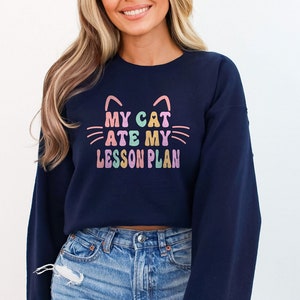 May include: A navy blue cropped sweatshirt with the text "My Cat Ate My Lesson Plan" in rainbow colors. The sweatshirt has a cat face graphic with pink ears and whiskers.