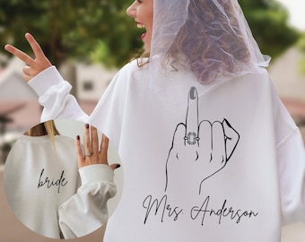 Könnte beinhalten: Weißer Pullover mit einem Design für eine zukünftige Braut. Die Rückseite des Pullovers zeigt eine Hand mit einem Ring am Mittelfinger, die den Mittelfinger zeigt, mit dem Text "Mrs. Anderson" darunter. Die Vorderseite des Pullovers trägt den Schriftzug "bride".