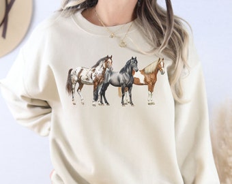 May include: A beige sweatshirt with a graphic print of three horses standing side by side. The horses are brown, black, and white.
