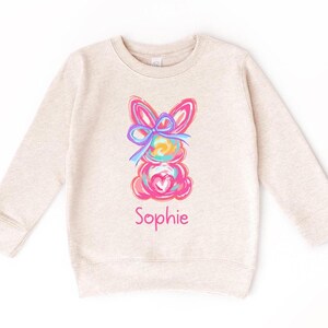 May include: A beige long-sleeved sweatshirt with a colorful bunny rabbit graphic and the name "Sophie" printed below it.