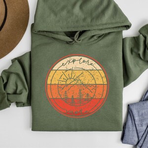 May include: A green hooded sweatshirt with a retro-style graphic of a mountain range, sun, and trees. The graphic is surrounded by a circle with the text "Explore More" in a vintage font.