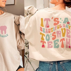 Peut inclure: Un sweat-shirt beige clair avec un motif de dent rose et le mot "Emily" en dessous. Le dos du sweat-shirt présente un motif coloré qui dit "C'est une belle journée pour sauver les dents".