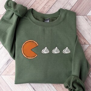 Puede incluir: Sudadera verde con un diseño de Pac-Man comiendo mini tartas de calabaza. El Pac-Man es naranja y las tartas son blancas con una corteza marrón.