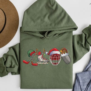May include: A green hooded sweatshirt with a Christmas-themed hockey design. The design features a hockey stick, a skate, a hockey helmet, and a puck, all decorated with Christmas lights and Santa hats.