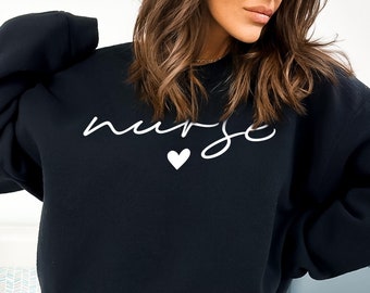 Könnte beinhalten: Schwarzer Pullover mit dem Wort "nurse" in weißer Kursivschrift und einem kleinen weißen Herz darunter.