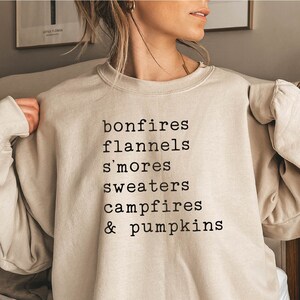 May include: A beige sweatshirt with the text "bonfires, flannels, s'mores, sweaters, campfires & pumpkins" printed in black.