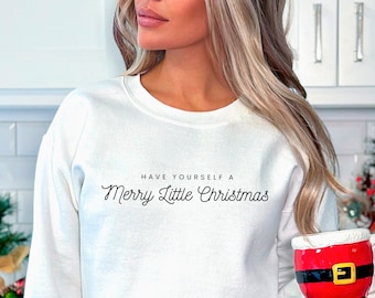 May include: White sweatshirt with the text "Have Yourself a Merry Little Christmas" in black script. The person is wearing blue jeans and holding a red Santa mug.