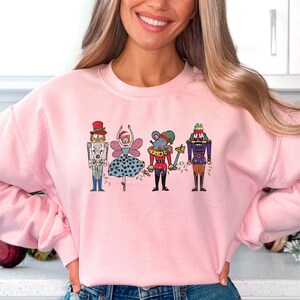 Può includere: Una felpa rosa con una grafica di una ballerina, uno schiaccianoci, un topo e un altro schiaccianoci. La grafica è in uno stile cartone animato colorato.