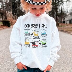 Peut inclure: Sweat-shirt blanc avec un graphique de véhicules de chantier et le texte "Dieu dit que je suis..." avec des versets bibliques. Le graphique comprend les mots "Jamais seul", "Victorieux", "Fort", "Digne", "Capable" et "Choisi".