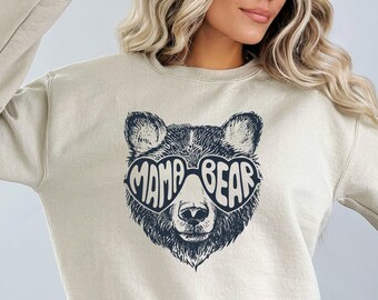 May include: Sand-colored sweatshirt featuring a black graphic of a bear's face wearing sunglasses. The sunglasses spell out "MAMA BEAR". The sweatshirt has a crew neck and long sleeves. The brand name "Zephyr Apparel" is printed on the upper right.