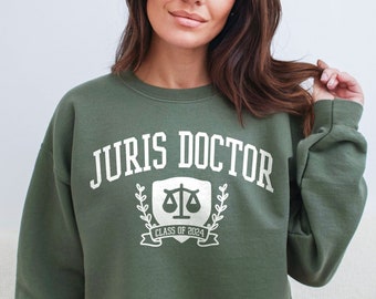 May include: A green sweatshirt with the text "JURIS DOCTOR" in white, a white scale of justice graphic, and the text "CLASS OF 2024" in white.