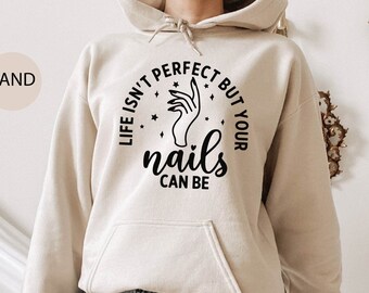 Op de afbeelding: Zandkleurige hoodie met een voorzak en een capuchon met trekkoord. De voorkant van de hoodie heeft de tekst "LIFE ISN'T PERFECT BUT YOUR nails CAN BE" in een cirkelvormig ontwerp met een handillustratie. De hoodie wordt gecombineerd met een spijkerbroekshort.