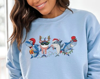 Puede incluir: Una sudadera de color azul claro con un gráfico navideño de cuatro tiburones de dibujos animados que llevan gorros de Papá Noel, cuernos de reno y un sombrero de copa. Los tiburones sostienen regalos y están rodeados de luces navideñas.