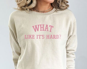 Könnte beinhalten: Ein beiger Pullover mit dem Aufdruck "WHAT LIKE IT'S HARD?" in Rosa.