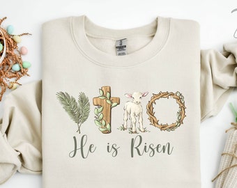 May include: A beige sweatshirt featuring a graphic design of a cross, a lamb, and a wreath. The text "He is Risen" is printed below the design.