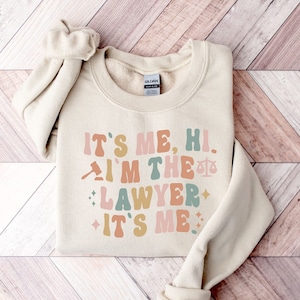 Peut inclure: Un sweat-shirt beige avec le texte "It's me, hi. I'm the lawyer. It's me." en lettres r&eacute;tro color&eacute;es. Un marteau et une balance de la justice sont inclus dans le design.