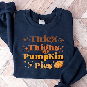 May include: Navy blue sweatshirt with the text "Thick Thighs Pumpkin Pies" in orange and brown. The design includes starburst and pie graphics. The sweatshirt is folded on a light wood grain surface.