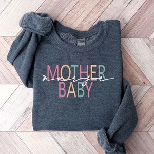 Puede incluir: Una sudadera gris oscura con el texto "MOTHER" y "BABY" en letras de colores. El texto est&aacute; en una fuente divertida y est&aacute; centrado en la sudadera.