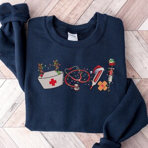 Puede incluir: Una sudadera azul marino con un dise&ntilde;o navide&ntilde;o que incluye un gorro de enfermera, un estetoscopio, un term&oacute;metro, una venda y una jeringa. El dise&ntilde;o est&aacute; formado por varios s&iacute;mbolos m&eacute;dicos decorados con luces de Navidad y un gorro de Pap&aacute; Noel.
