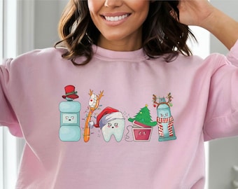 May include: Pink sweatshirt featuring a Christmas-themed dental hygiene design. The design includes a toothbrush, toothpaste, mouthwash, floss, and a tooth wearing a Santa hat. The text "Merry" is written in a festive font.