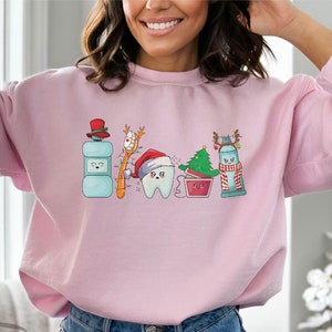 以下が含まれることがあります： クリスマスをテーマにした歯の衛生のデザインが施されたピンク色のスウェットシャツ。デザインには、歯ブラシ、歯磨き粉、マウスウォッシュ、フロス、サンタの帽子をかぶった歯が描かれています。テキストの「メリー」は、お祝いのフォントで書かれています。