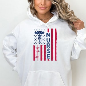 Puede incluir: Sudadera con capucha blanca con un diseño de bandera estadounidense roja, blanca y azul. La bandera tiene un símbolo de caduceo médico y la palabra "NURSE" impresa verticalmente en el lado derecho.