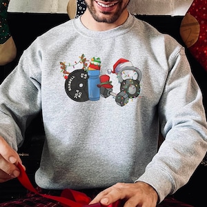 K&ouml;nnte beinhalten: Ein graues Sweatshirt mit einem weihnachtlichen Design, das eine Langhantel, Hanteln, eine Kettlebell, eine Wasserflasche und eine Weihnachtsm&uuml;tze zeigt. Das Design enth&auml;lt Weihnachtslichter und ein Rentier.