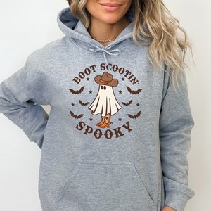 Peut inclure: Sweat-shirt gris à capuche avec un motif de fantôme portant un chapeau de cowboy et des bottes. Le texte "Boot Scootin' Spooky" est imprimé dans une police de caractères de style vintage.