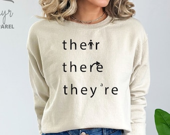 Op de afbeelding: Een beige sweatshirt met de woorden "their", "there" en "they're" erop gedrukt. Het woord "there" heeft een pijl die naar rechts wijst.