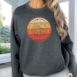 May include: A heather dark grey sweatshirt with a vintage-style graphic of a mountain range, sun, and trees. The graphic is surrounded by a red, orange, and yellow striped circle. The text "explore more" is written in a handwritten font above the graphic.