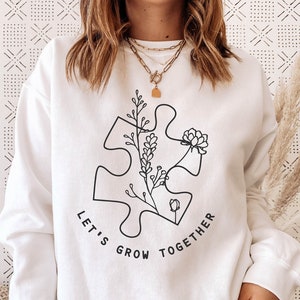 May include: White sweatshirt with a black outline of a puzzle piece with flowers growing inside. The text "Let's Grow Together" is written around the puzzle piece.