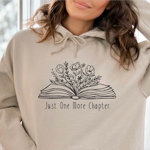 Peut inclure: Un sweat &agrave; capuche beige avec un motif noir d'un livre ouvert avec des fleurs qui en sortent. Le texte "Just One More Chapter" est imprim&eacute; sous le livre.